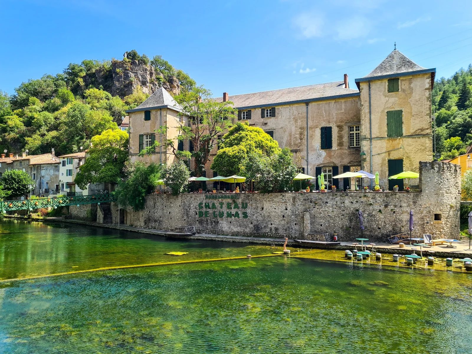 Vue extérieure du Château de Lunas depuis la rivière avec terrasse et parasols verts, lieu de mariage en Hérault