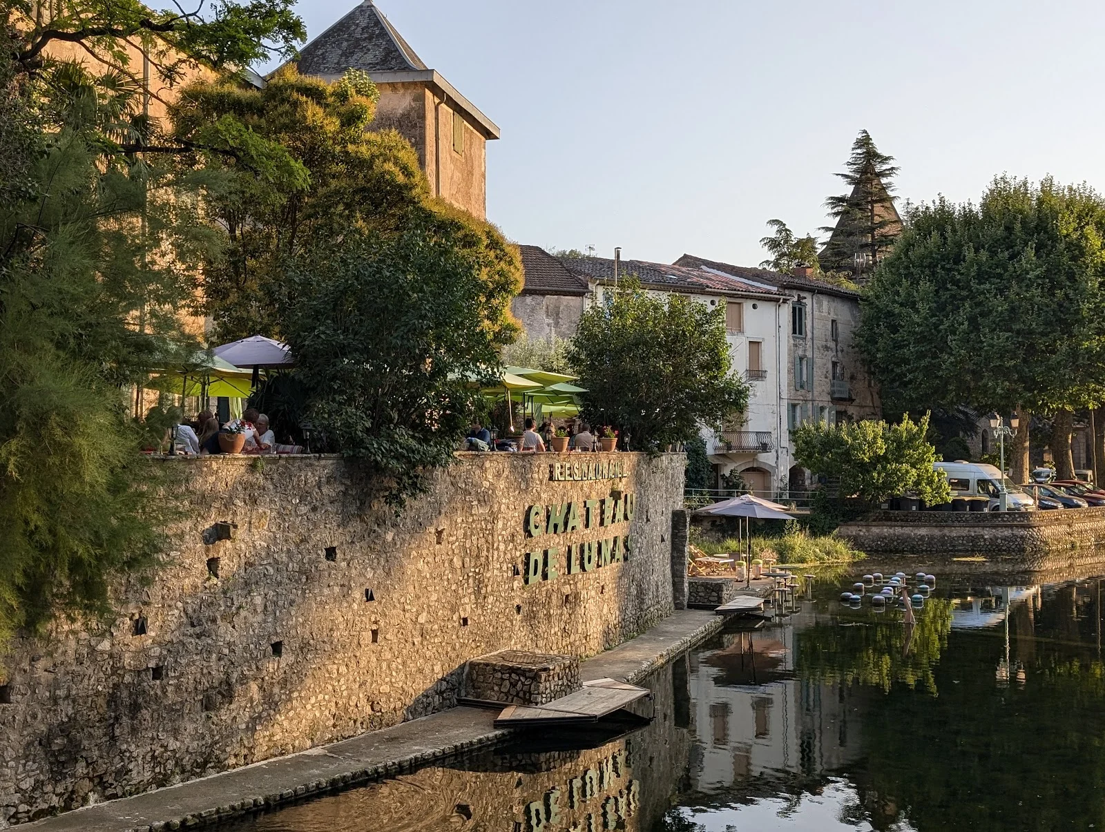 Vue en soirée du Château de Lunas depuis la rivière avec lumière dorée et terrasse animée, Hérault
