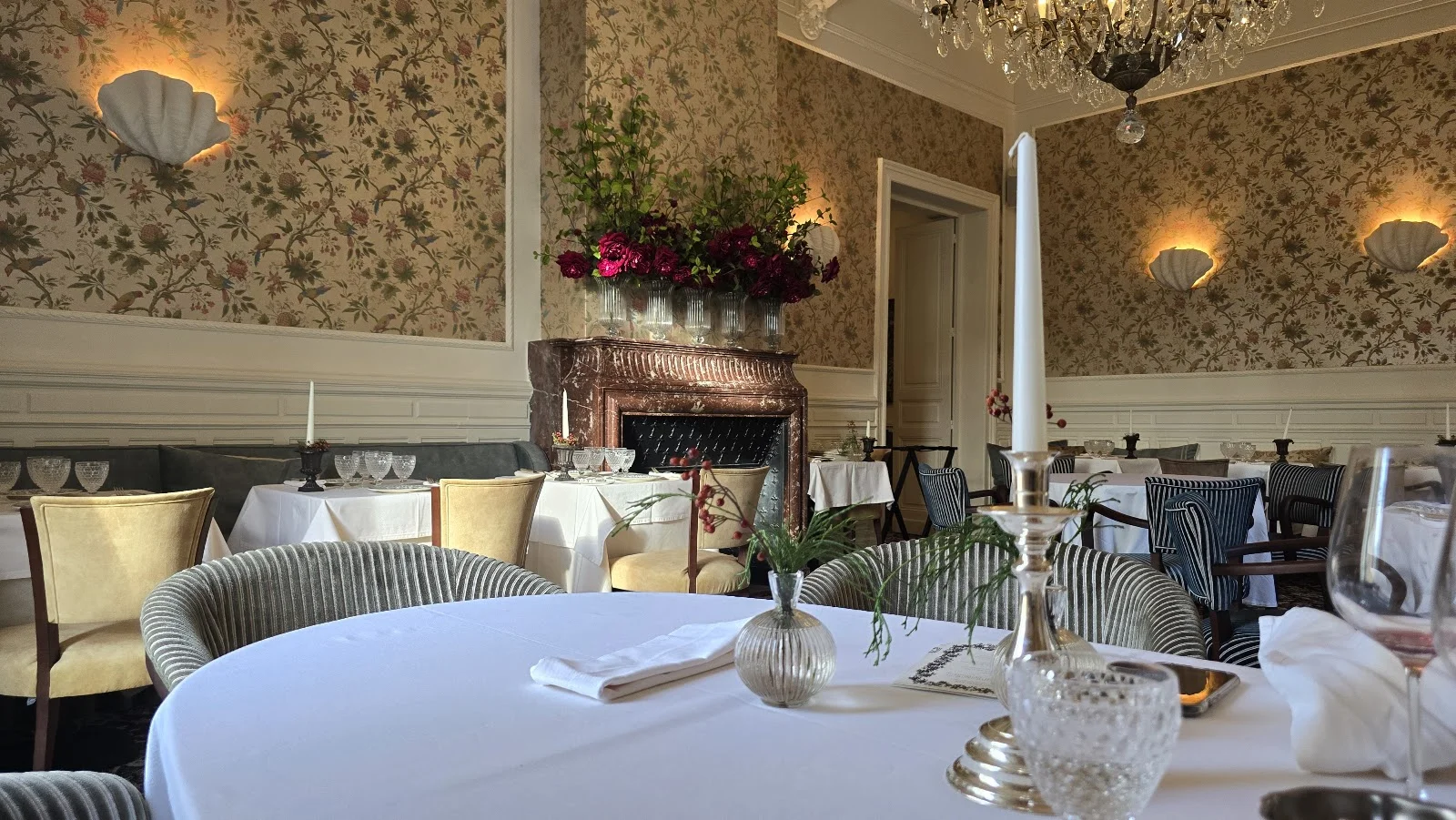 Salle de réception lumineuse du Château Paloma dans l'Hérault avec cheminée fleurie, lustres cristal et tables dressées pour mariage