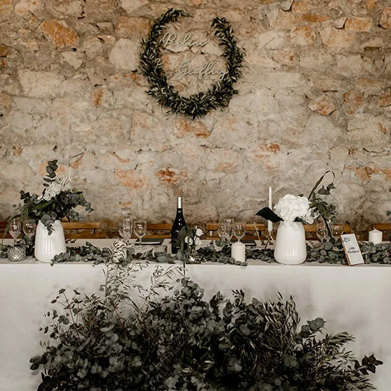 Table d'honneur de mariage décorée d'eucalyptus et fleurs blanches devant un mur en pierre au Domaine de Fangouse Hérault