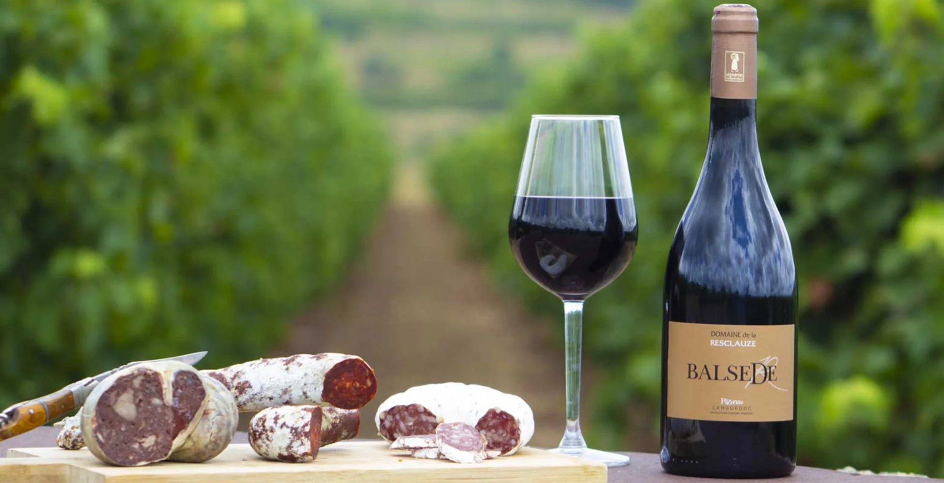 Bouteille de vin Balsède du Domaine de la Resclauze avec verre de rouge et saucissons devant les vignes de l'Hérault
