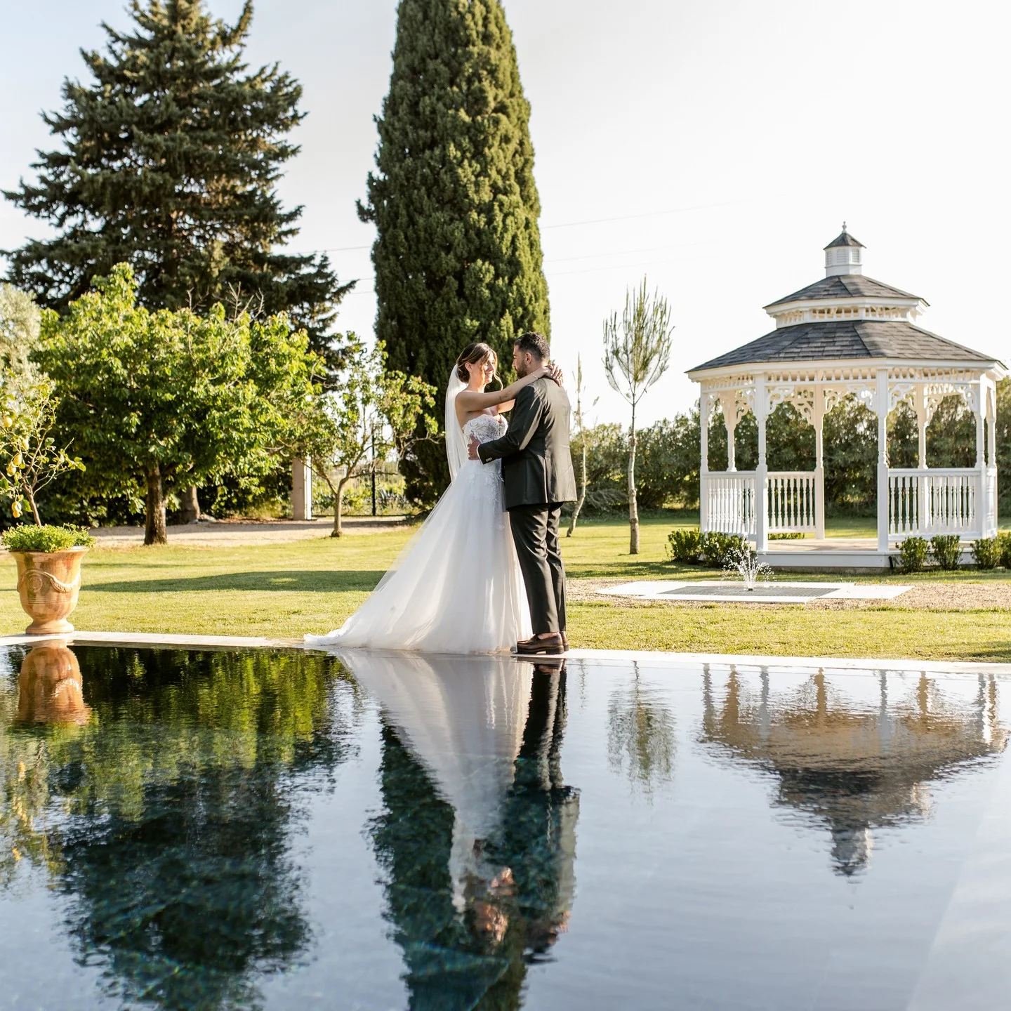 Couple de mariés enlacés au bord du miroir d'eau avec la bastide et le kiosque du Domaine de la Trinité, Hérault
