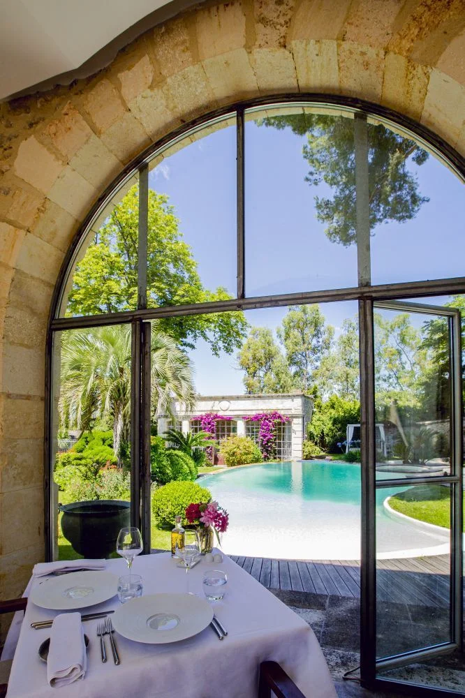 Table de restaurant dressée face à une baie vitrée arquée avec vue sur la piscine et le jardin du Domaine de Verchant, Hérault