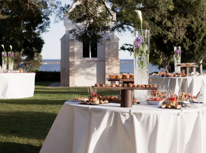 Buffet cocktail de mariage avec mignardises et vase fleuri, chapelle et étang en fond au Domaine des Moures, Hérault