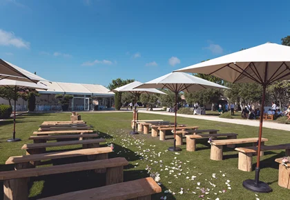 Espace cocktail en plein air au Domaine des Moures avec tables bois, bancs rustiques et parasols blancs, Hérault