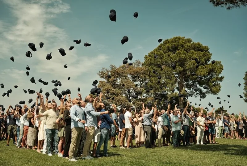 Foule joyeuse lançant des casquettes en l'air, grand parc arboré, ambiance festive Domaine du Mas Neuf Vic-la-Gardiole mariage Hérault Occitanie