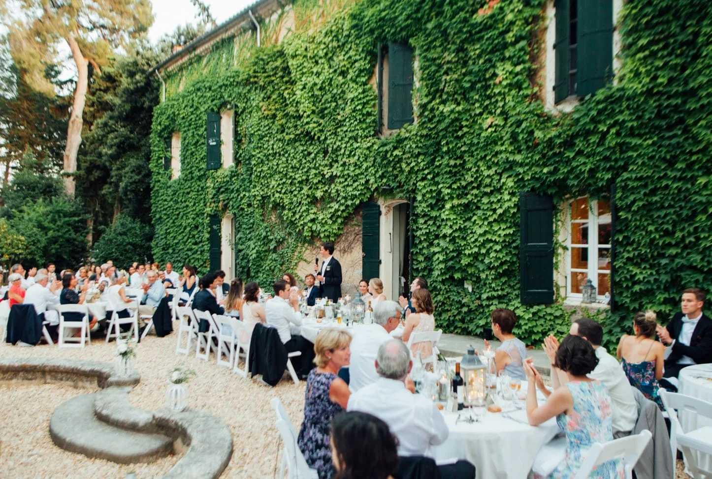 Dîner de mariage en extérieur devant la façade en lierre du Domaine Saint-Hilaire dans l'Hérault avec discours des mariés