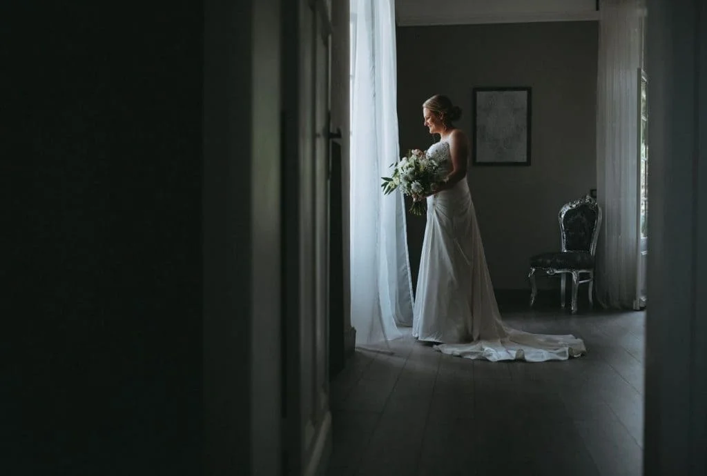 Mariée en robe blanche avec bouquet regardant par la fenêtre dans la chambre du Domaine Saint Hilaire en Hérault