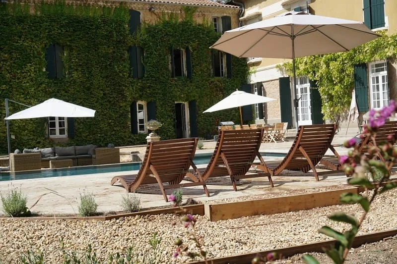 Piscine du Domaine Saint Hilaire en Hérault avec transats en bois et parasols blancs face au mas provençal