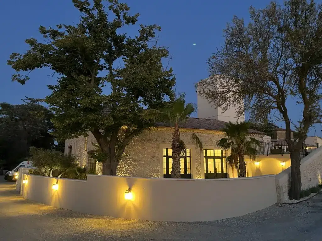 Façade extérieure des Granges Saint Paul illuminée de nuit avec murs en pierre et palmiers, Hérault