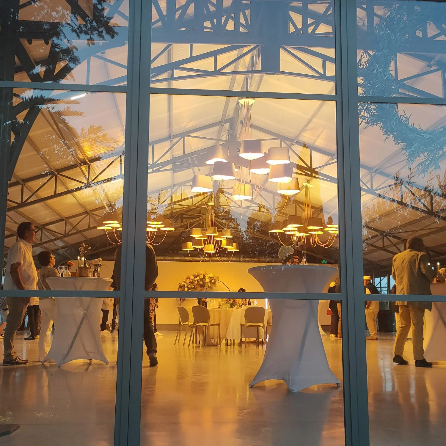 Salle de réception du Mas Aubrilha illuminée la nuit vue à travers les baies vitrées, ambiance dorée cocktail mariage Hérault