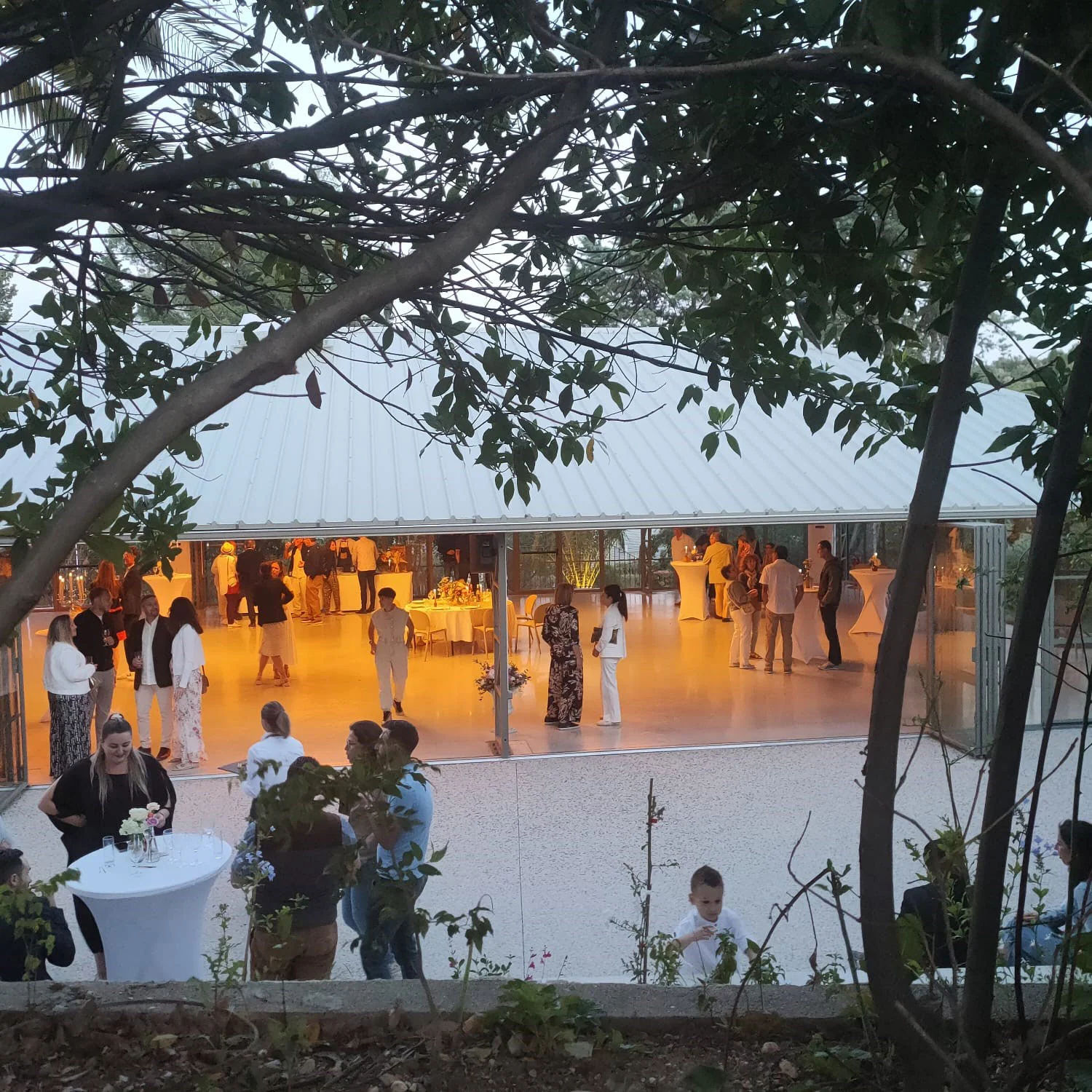 Soirée mariage au Mas Aubrilha, invités entre la salle vitrée illuminée et le jardin nocturne, domaine Hérault