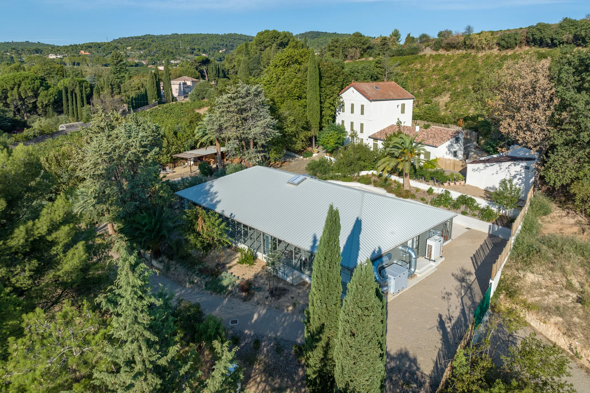 Vue aérienne du Mas Aubrilha en Hérault montrant la salle de réception vitrée et la bastide blanche entourées de pins