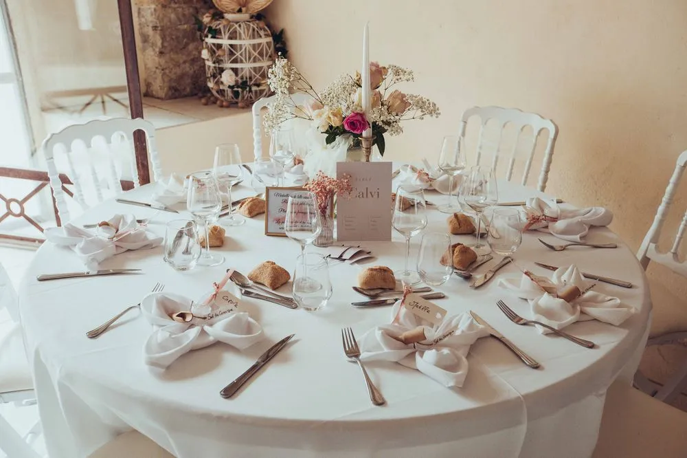 Table ronde dressée pour un mariage avec vaisselle, verres, serviettes pliées et centre de table floral — Camille Réceptions, traiteur de mariage à Clermont-l’Hérault