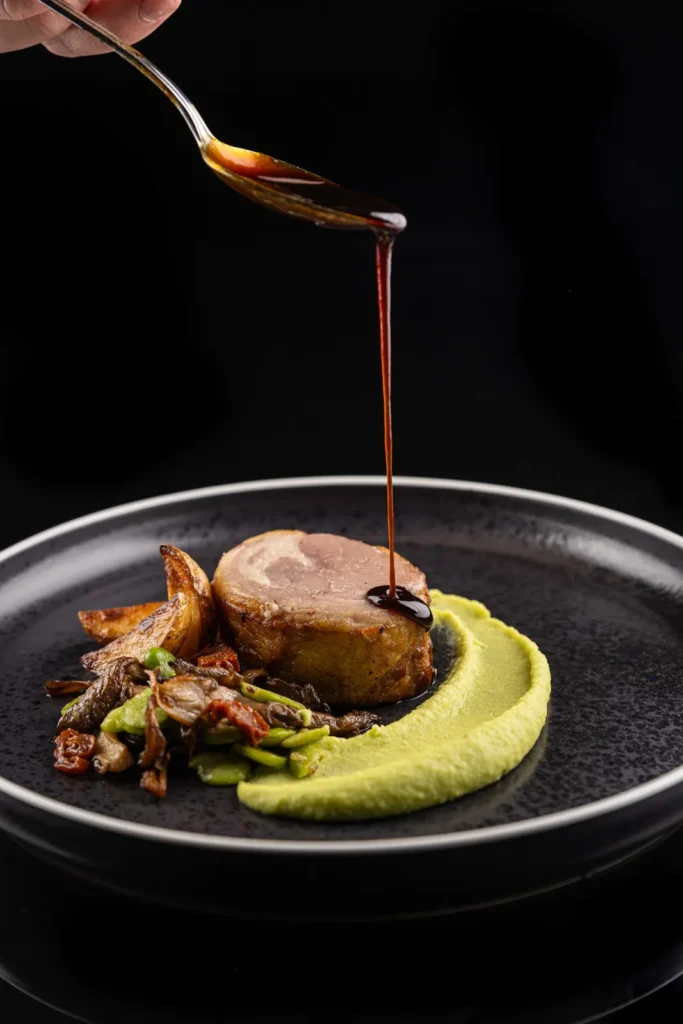 Main versant une sauce brune sur un plat de viande roulée, purée verte et légumes rôtis — Traiteur L’Évidence, Lattes