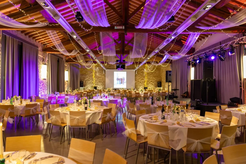 Grande salle de réception de mariage avec tables rondes dressées, chaises décorées et éclairage violet — Andrieux Traiteur, Lavérune