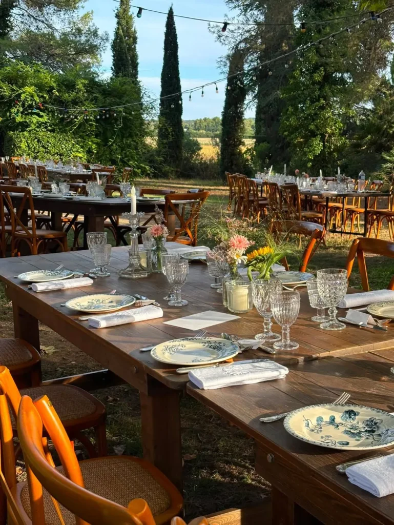 Longues tables en bois dressées pour un mariage en extérieur, vaisselle et décorations florales sous les arbres — Andrieux Traiteur, Lavérune