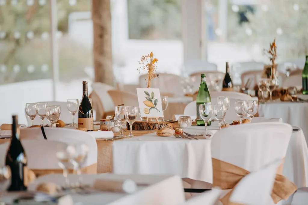 Tables rondes dressées pour un mariage, nappes blanches, chemin de table en jute, bouteilles de vin et décoration citron — Traiteur d’Amélie, Mauguio