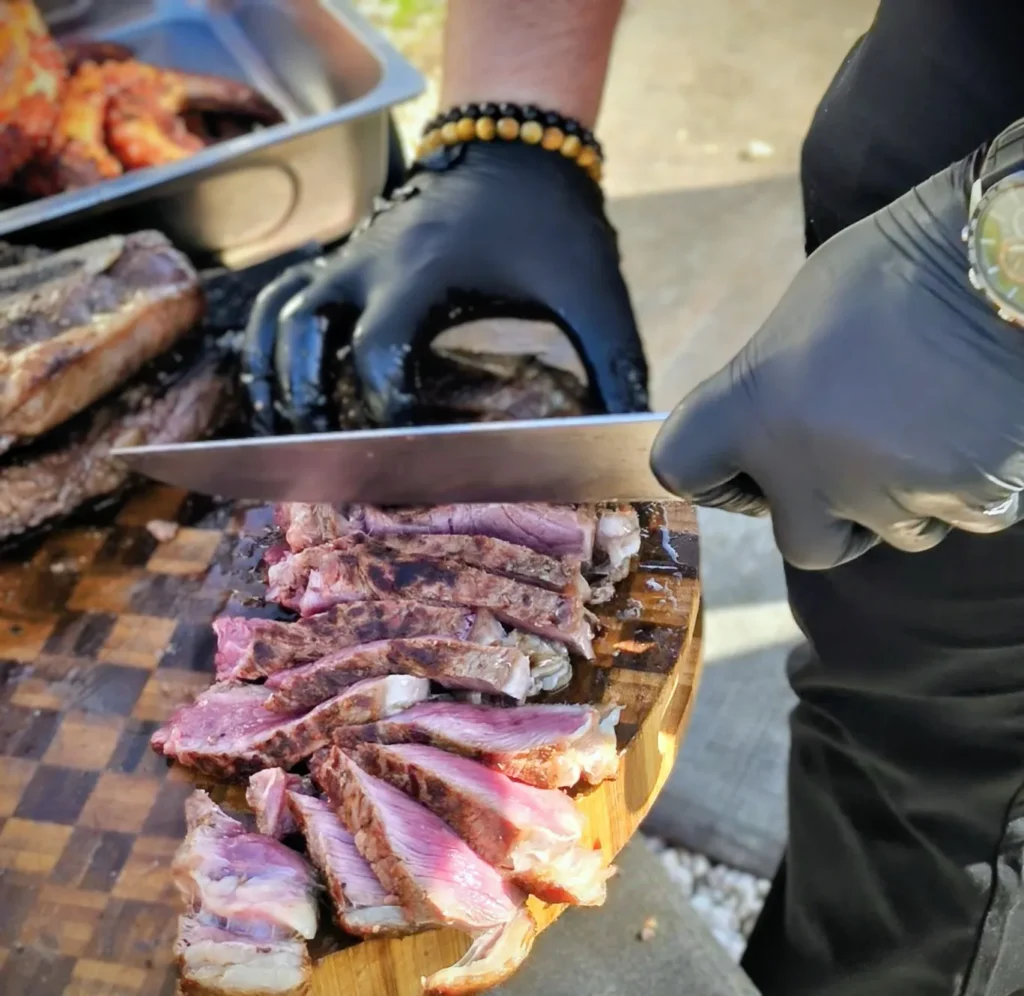 Un chef en gants noirs découpe une pièce de viande grillée saignante sur une planche en bois — Brasero Gourmand, traiteur de mariage à Montpellier