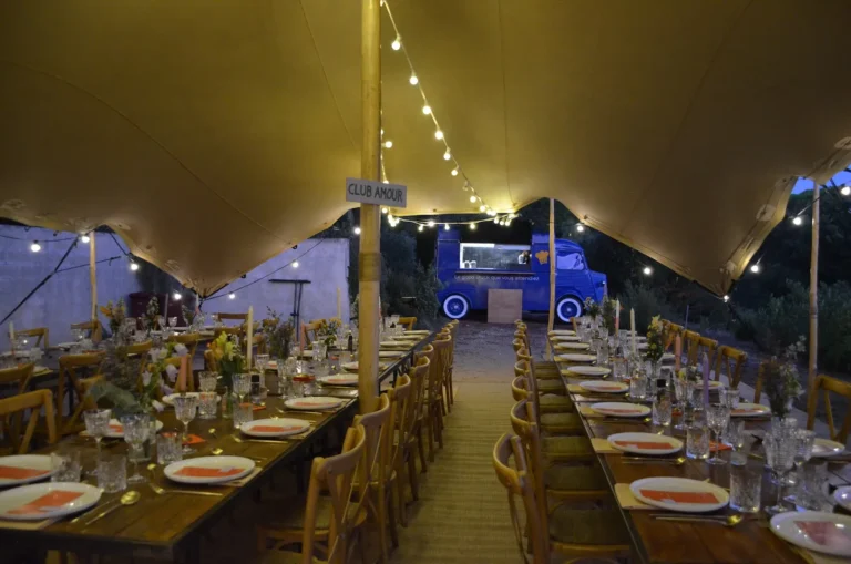 Longues tables en bois dressées sous une tente stretch illuminée par des guirlandes, avec un food truck bleu en arrière-plan — Général Moutarde, traiteur de mariage à Montpellier