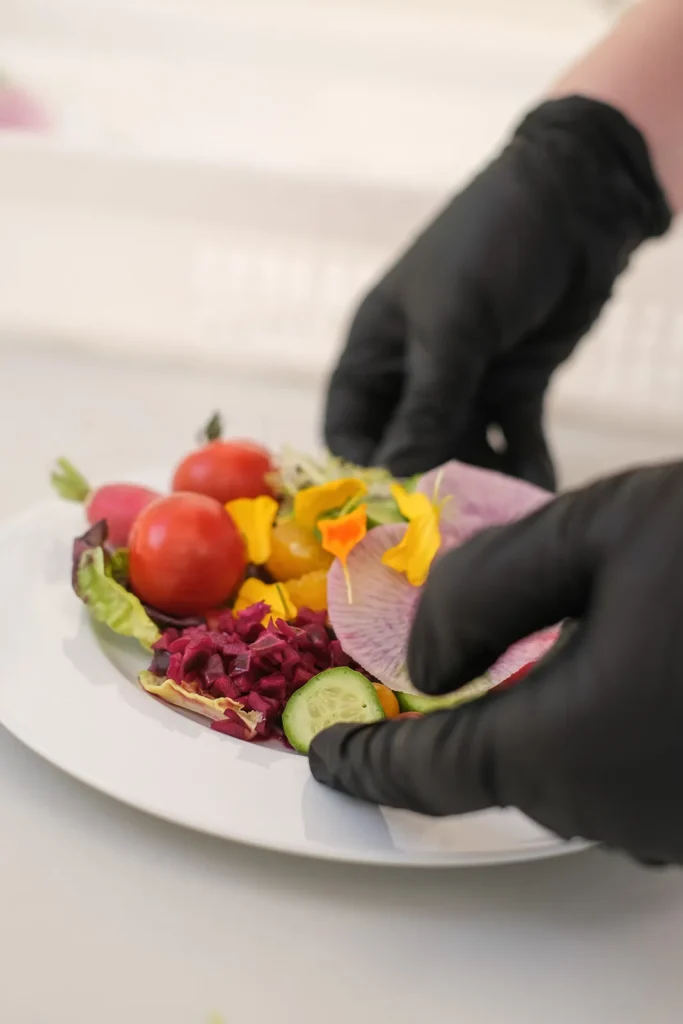 Mains gantées disposant légumes frais et fleurs comestibles sur une assiette blanche — Audrey Traiteur et Saveurs, mariages à Murviel-lès-Béziers