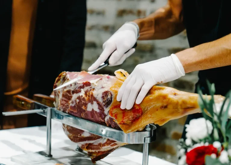 Mains gantées d’un traiteur découpant finement un jambon cru sur un support en acier — L’Occitane Traiteur, mariages à Nîmes