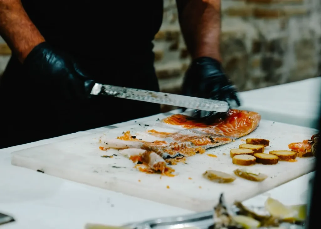 Mains d’un chef en gants noirs tranchant un filet de saumon frais — L’Occitane Traiteur, Nîmes